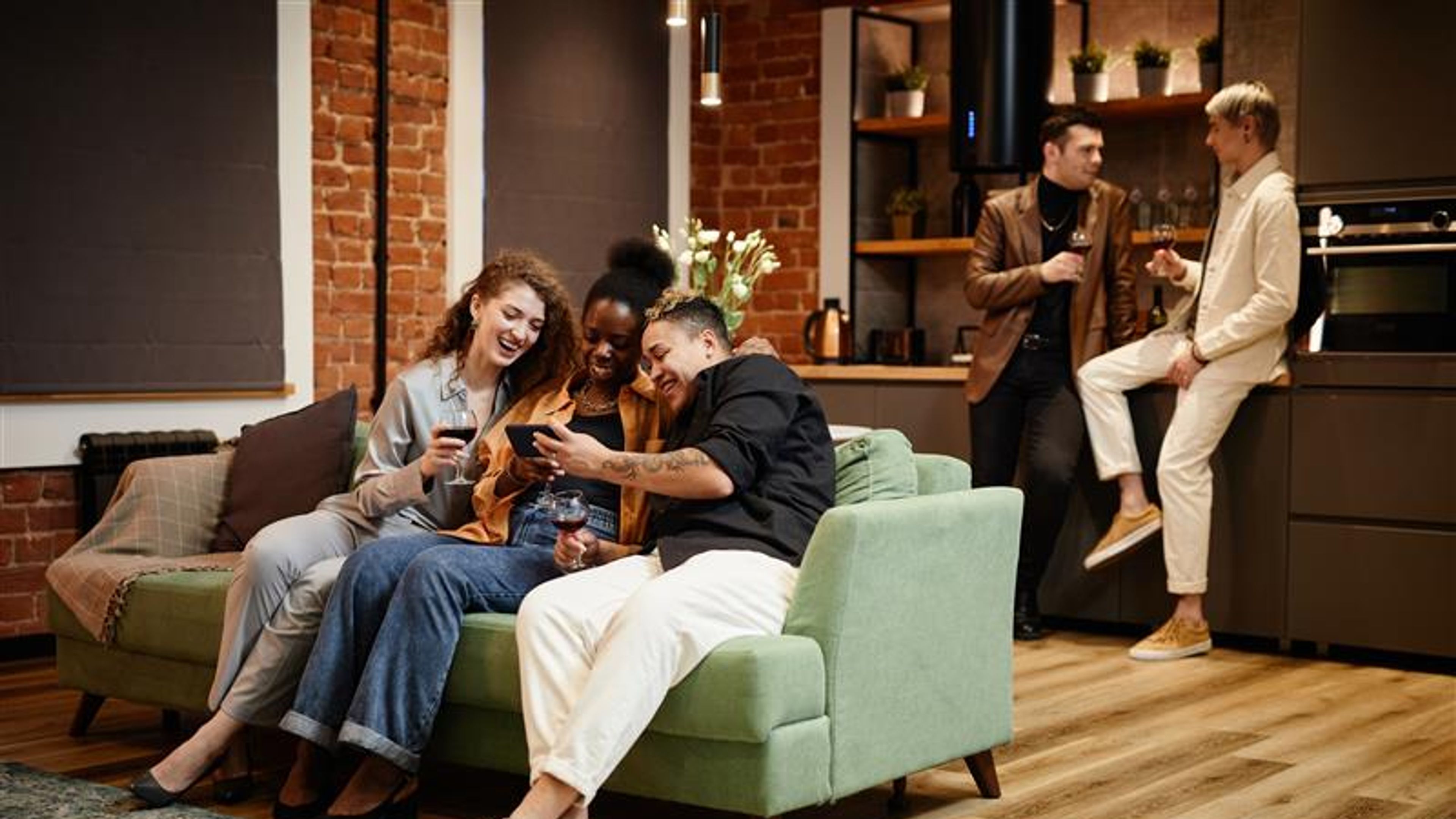 Three happy intercultural girls are looking through photos on their smartphones while resting on a couch in the kitchen. 