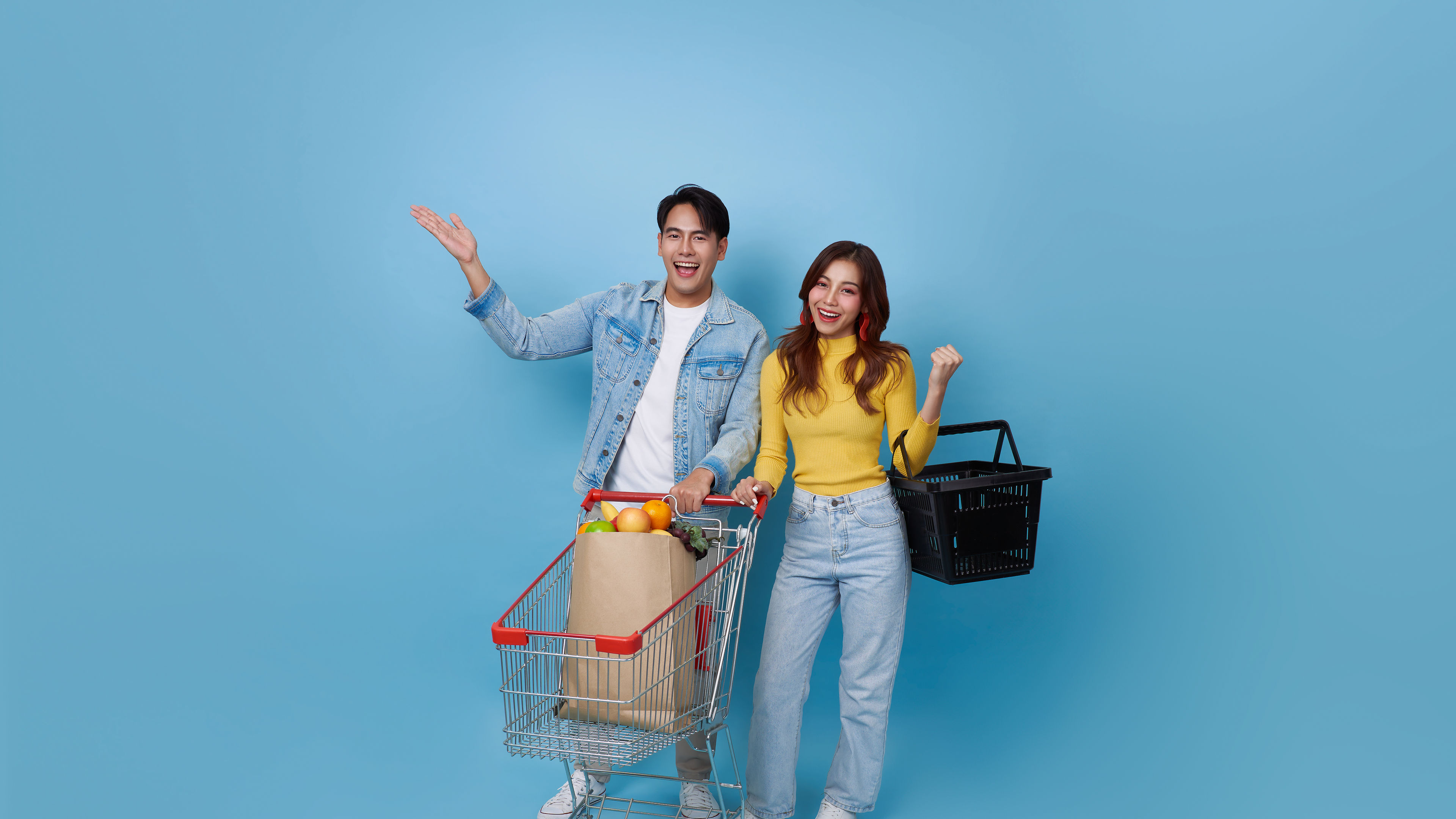 Young cheerful Asian couple with shopping trolley cart and holding shopping bag in a blue background.