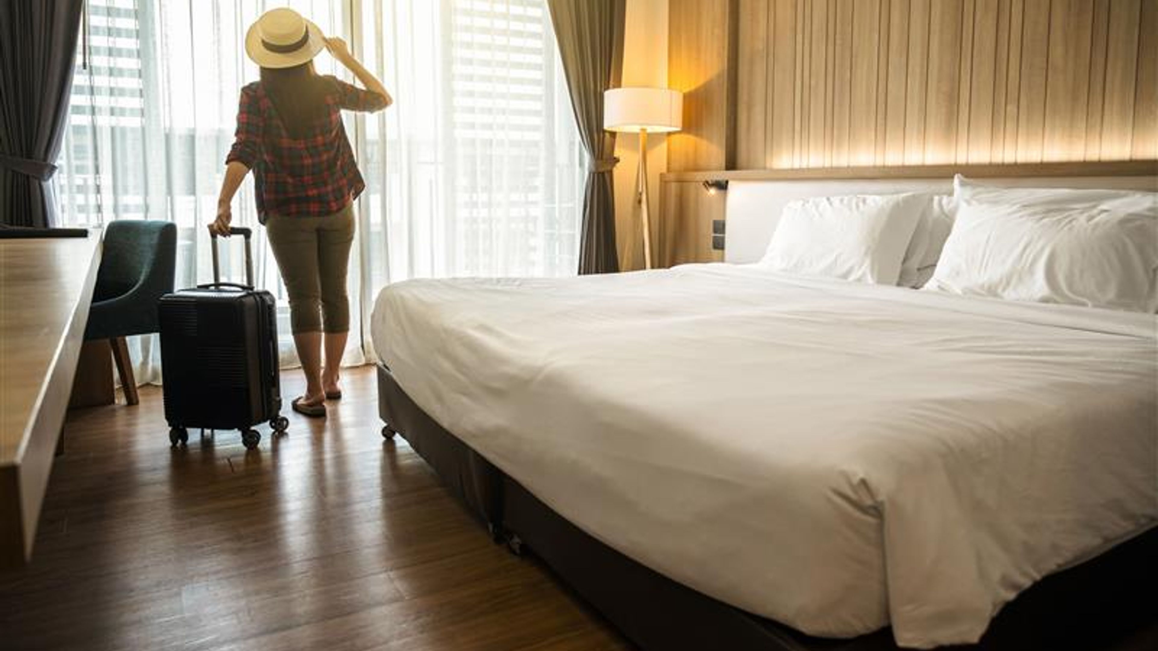 Một người phụ nữ đứng bên cửa sổ phòng khách sạn với hành lý bên cạnh, mỉm cười khi vui vẻ ngắm nhìn khung cảnh tuyệt đẹp bên ngoài