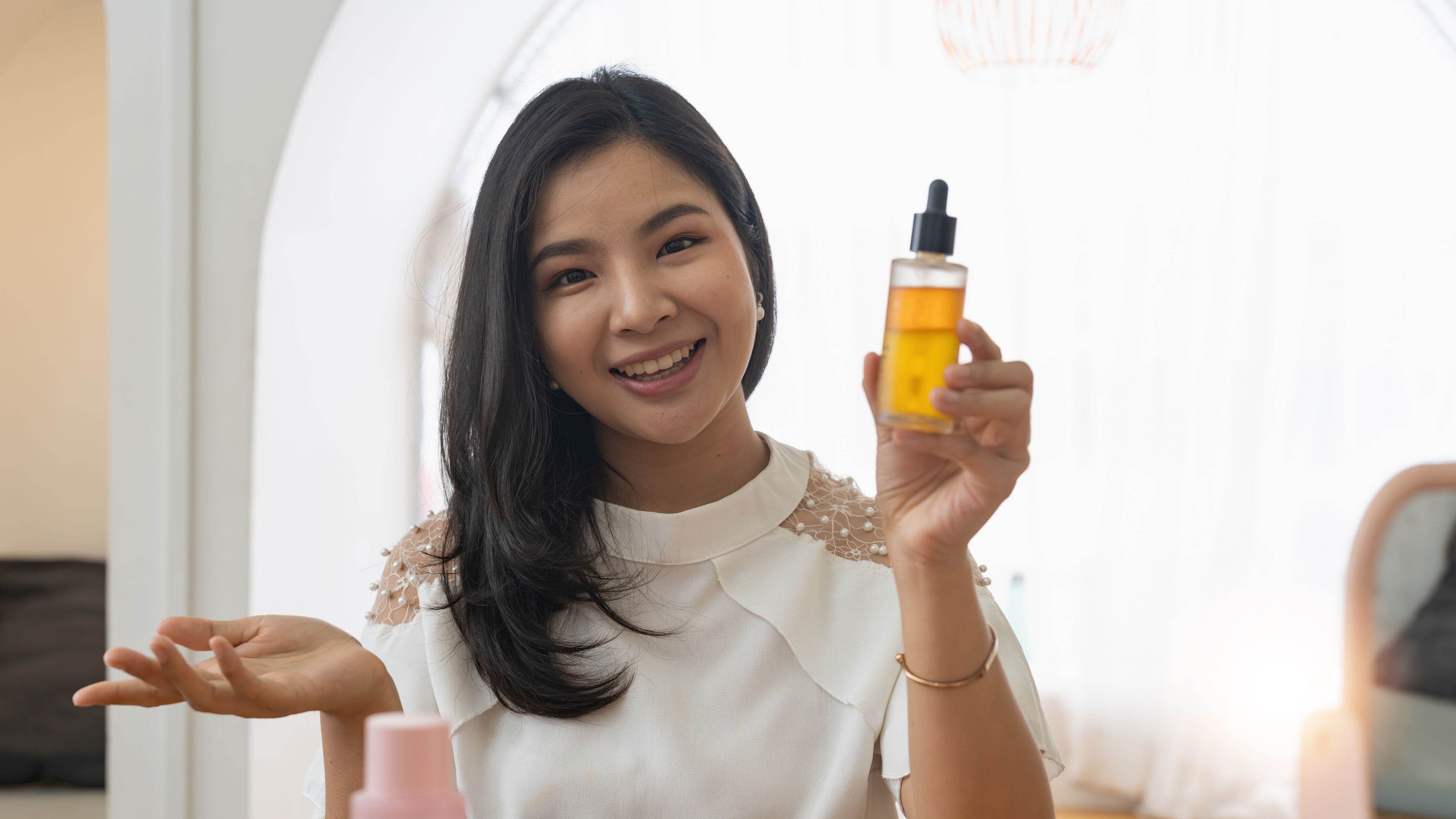 Smiling woman holding a skincare serum bottle while sitting at a table with beauty products.