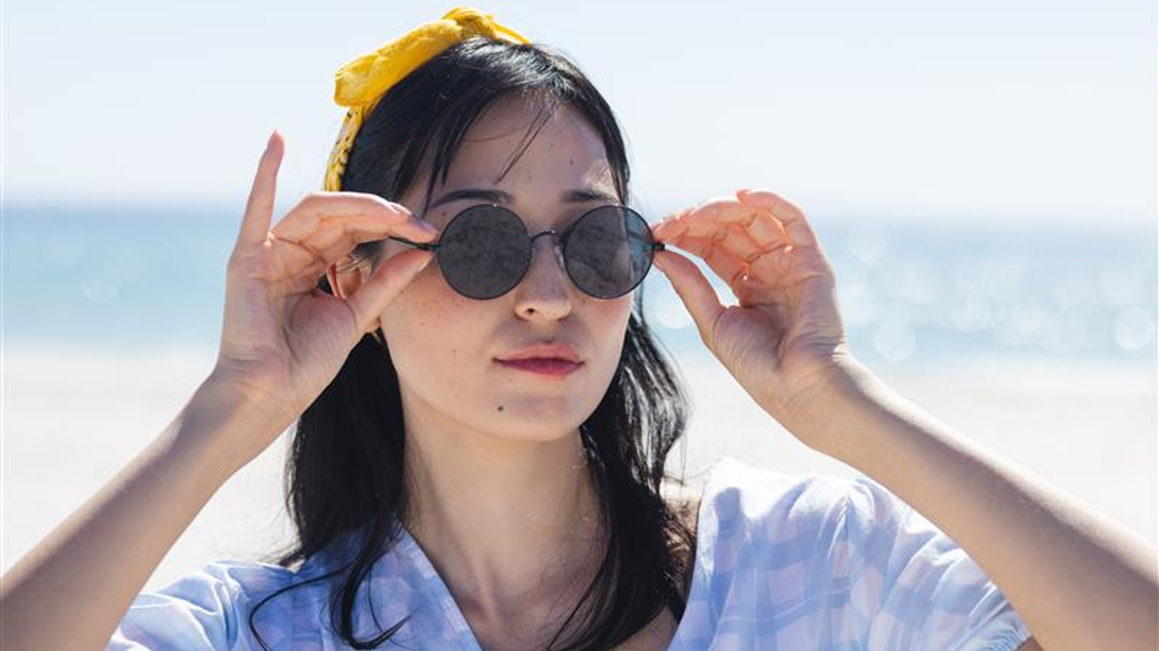 A young woman adjusts her sunglasses at the beach, unaltered. Her relaxed demeanor suggests a leisurely day outdoors. 