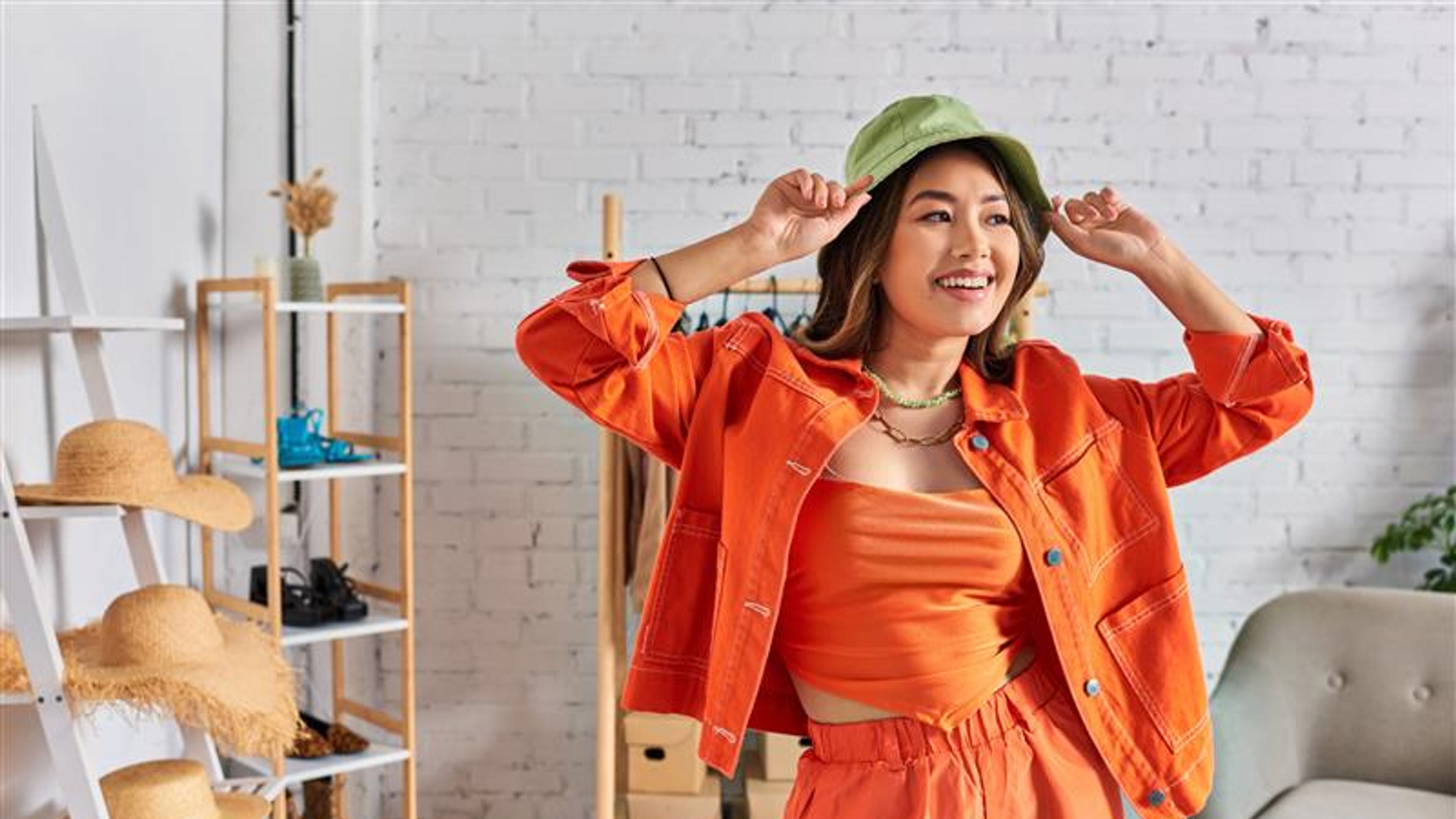 Delighted asian designer in orange clothes wearing panama hat and looking away in own atelier 