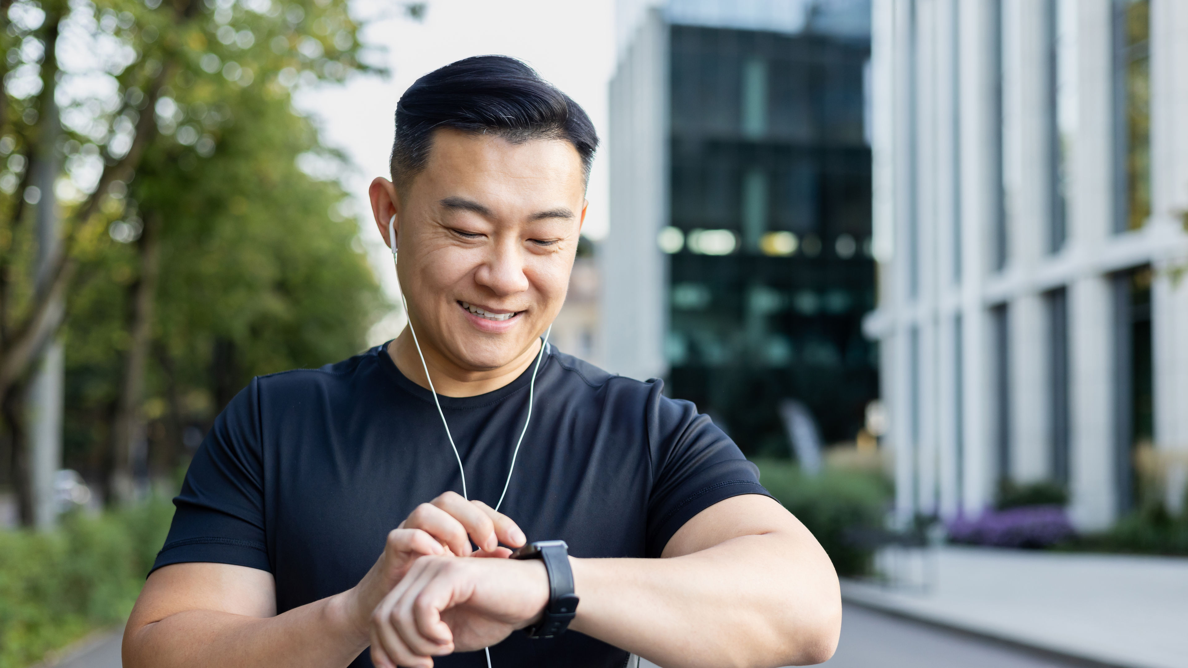 Young man standing on city street wearing headphones and looking at smart watch 