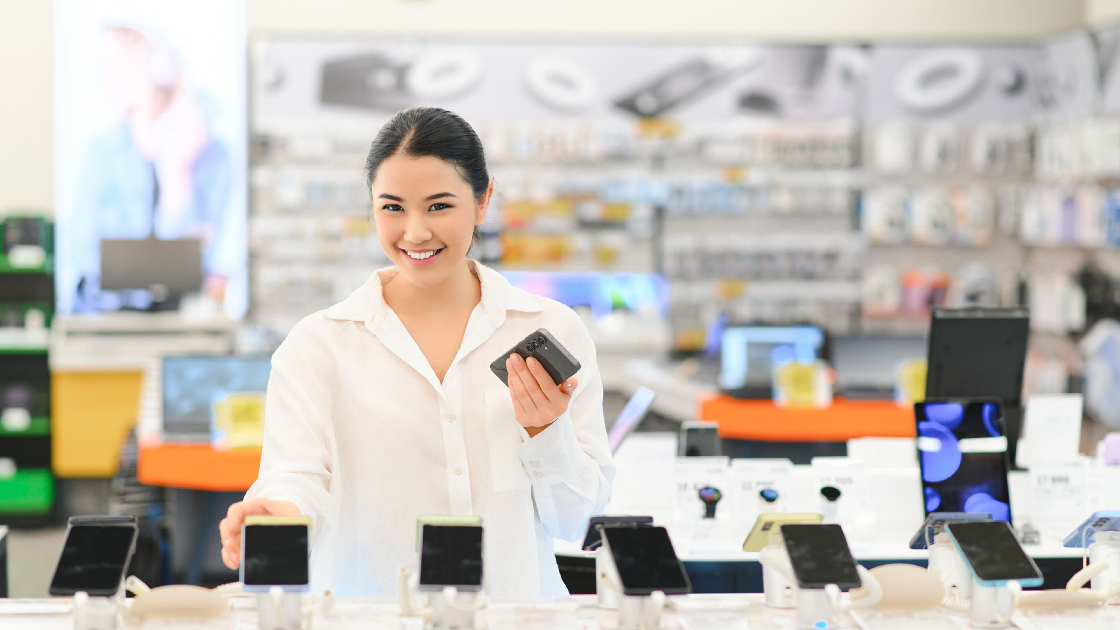 An Asian girl in white dress browsing smartphones in a tech store posing the picture with a smiling face.