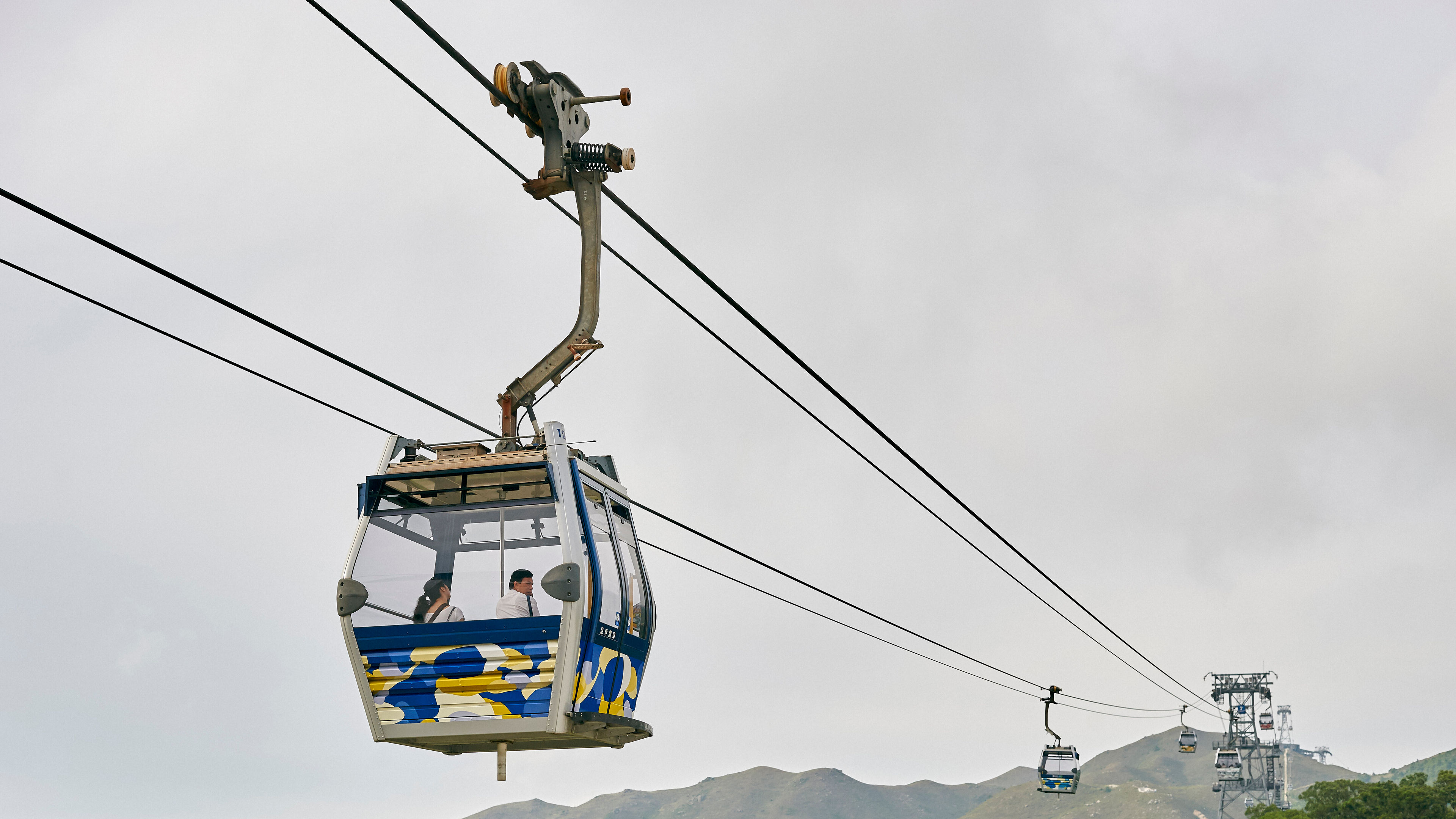 People are riding in a cable car above green hills
