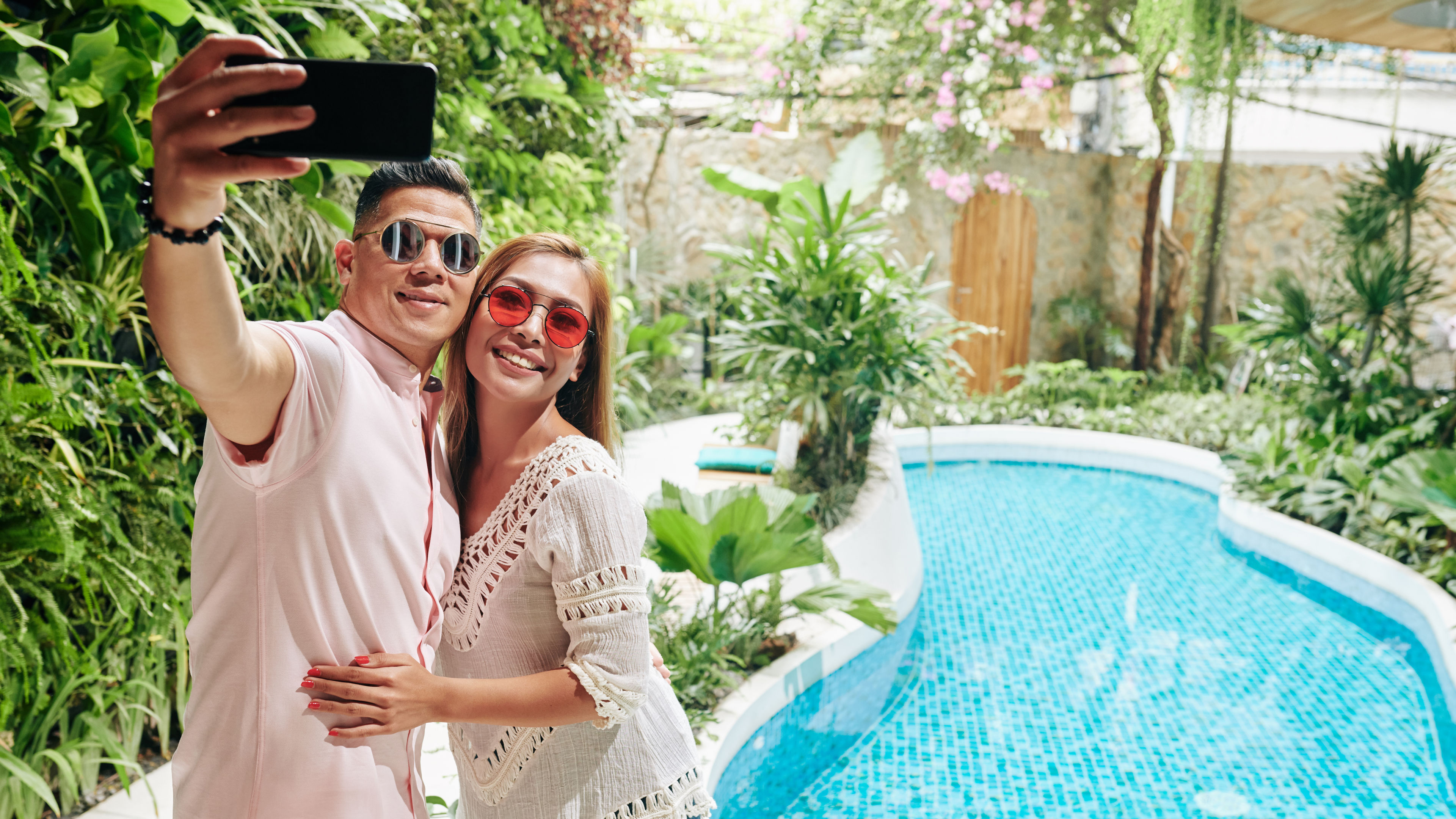 A couple in sunglasses takes a selfie by a blue tiled pool surrounded by tropical plants and flowers.