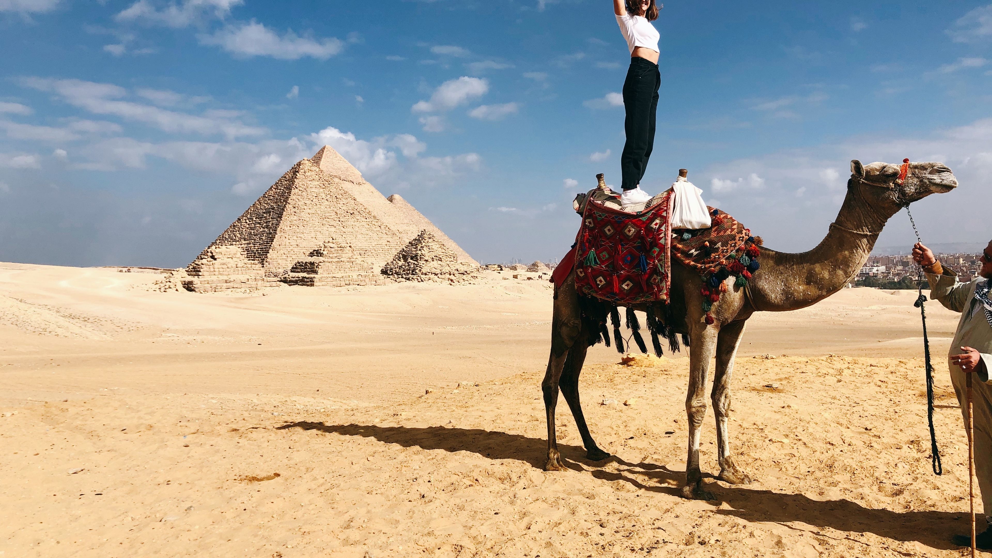 A girl standing on top of a camel with the pyramids visible in the background.