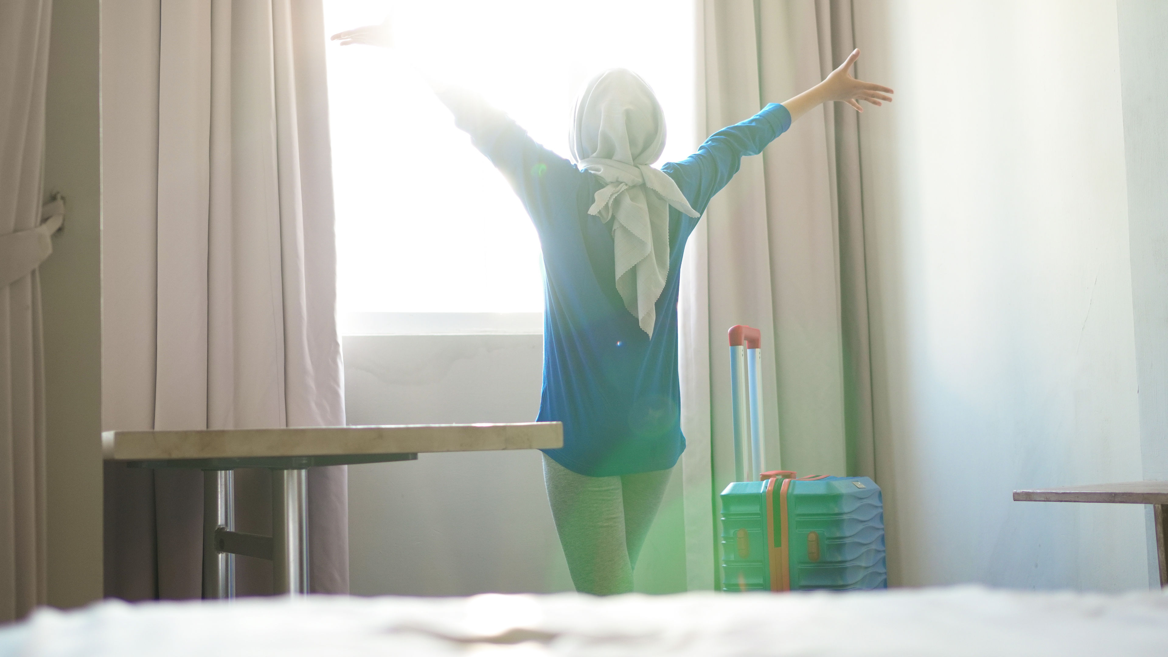 A person facing a sunlit window with arms outstretched, a teal suitcase placed nearby in the room.