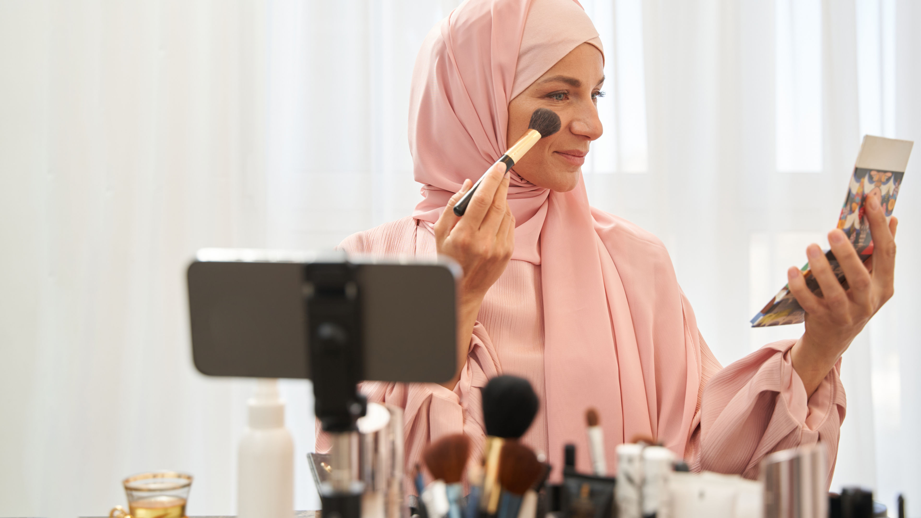 A woman applying makeup to her face with a focused expression, while various skincare essentials are neatly arranged on the table in front of her.