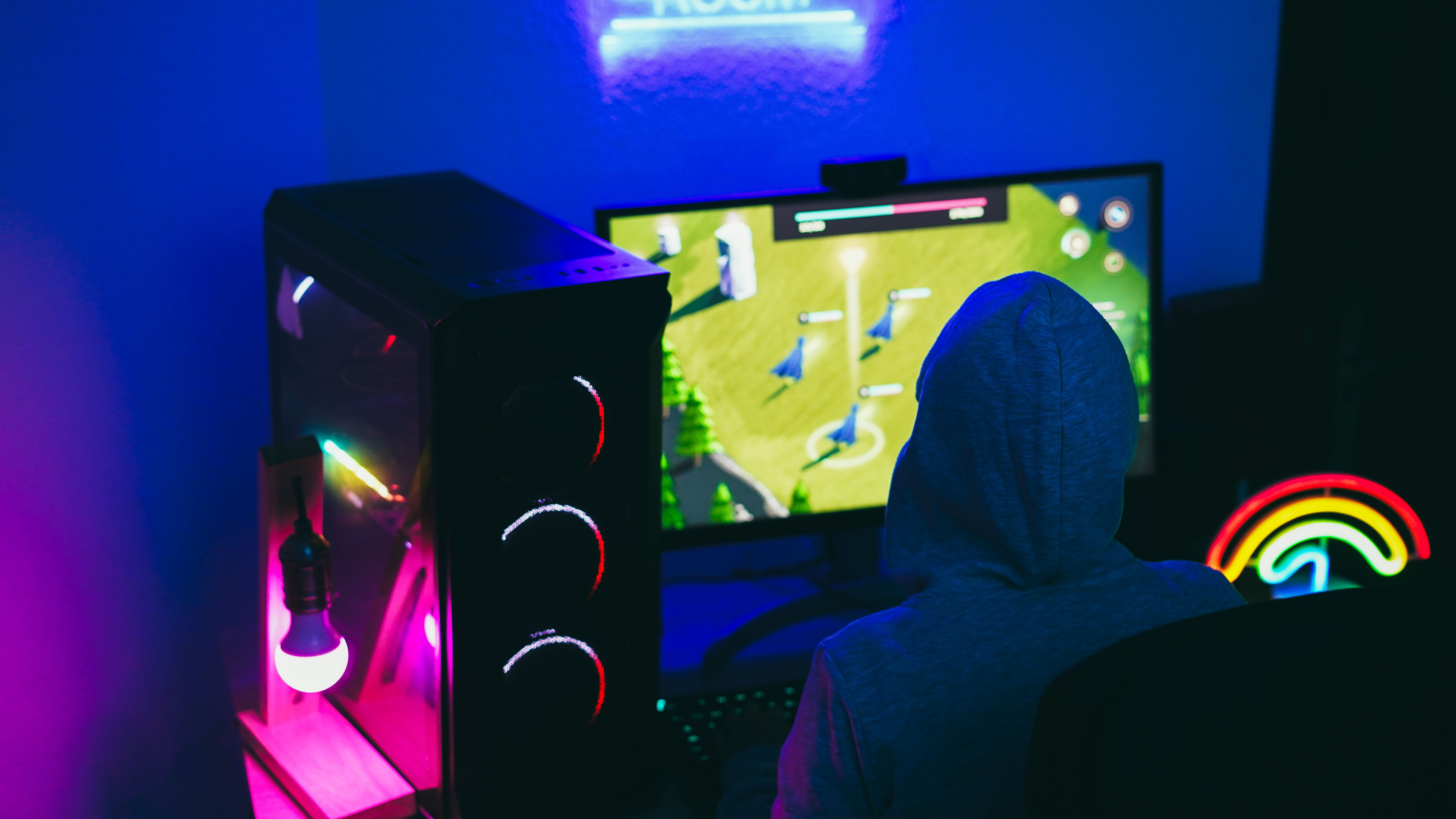A person in a hoodie playing a strategy game on a PC in a neon-lit gaming setup.