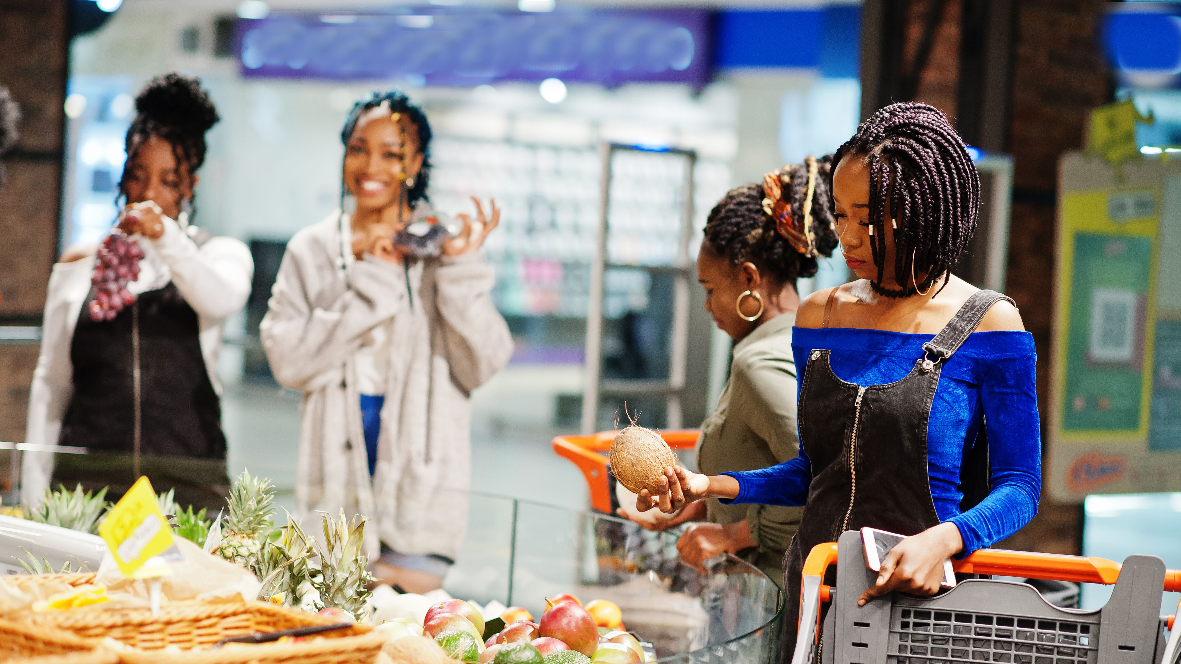 Four women shopping for fresh groceries at a market, with one holding a coconut.