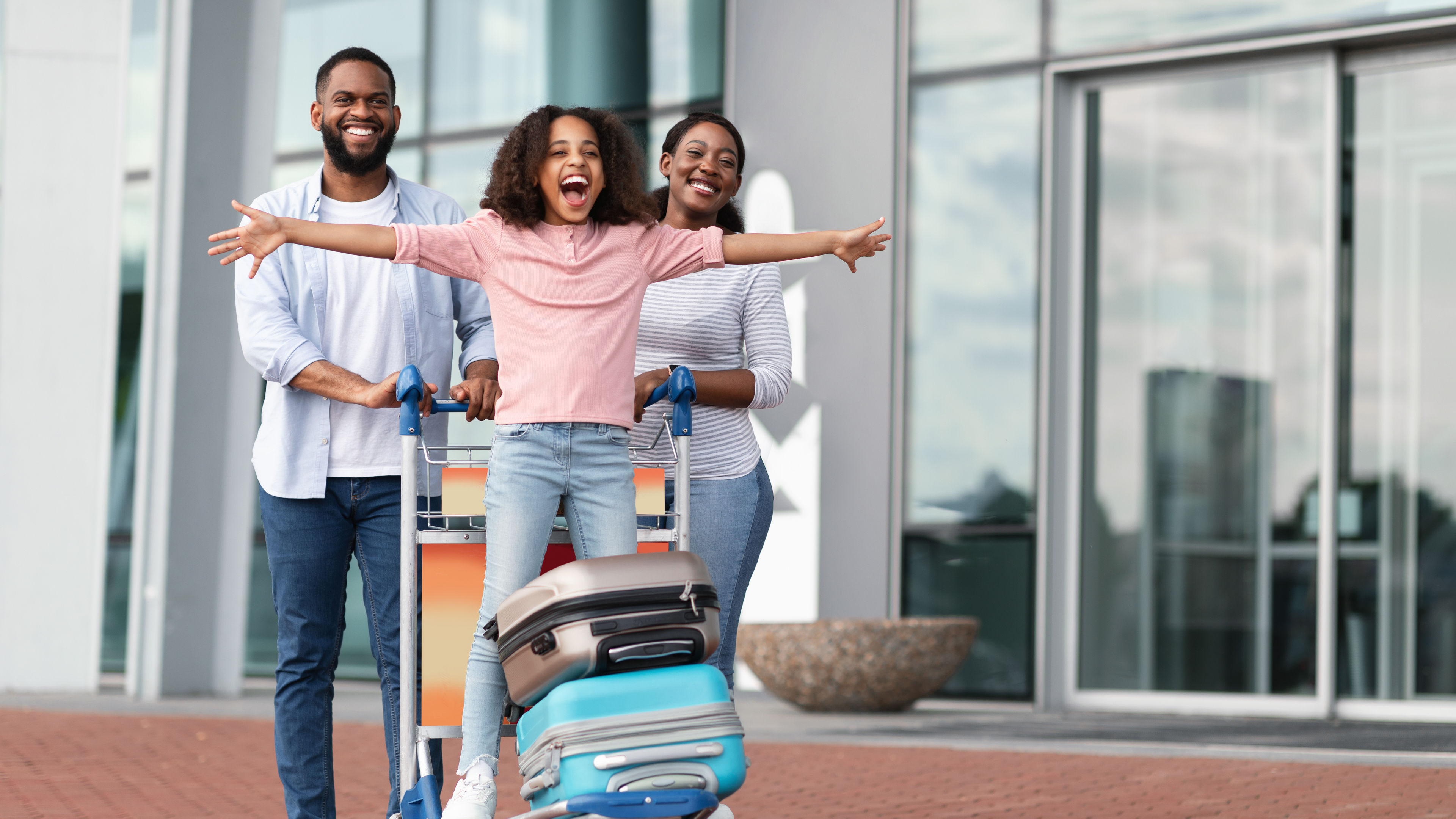 A joyful Black family preparing for a trip, with a child playfully standing on a luggage trolley.
