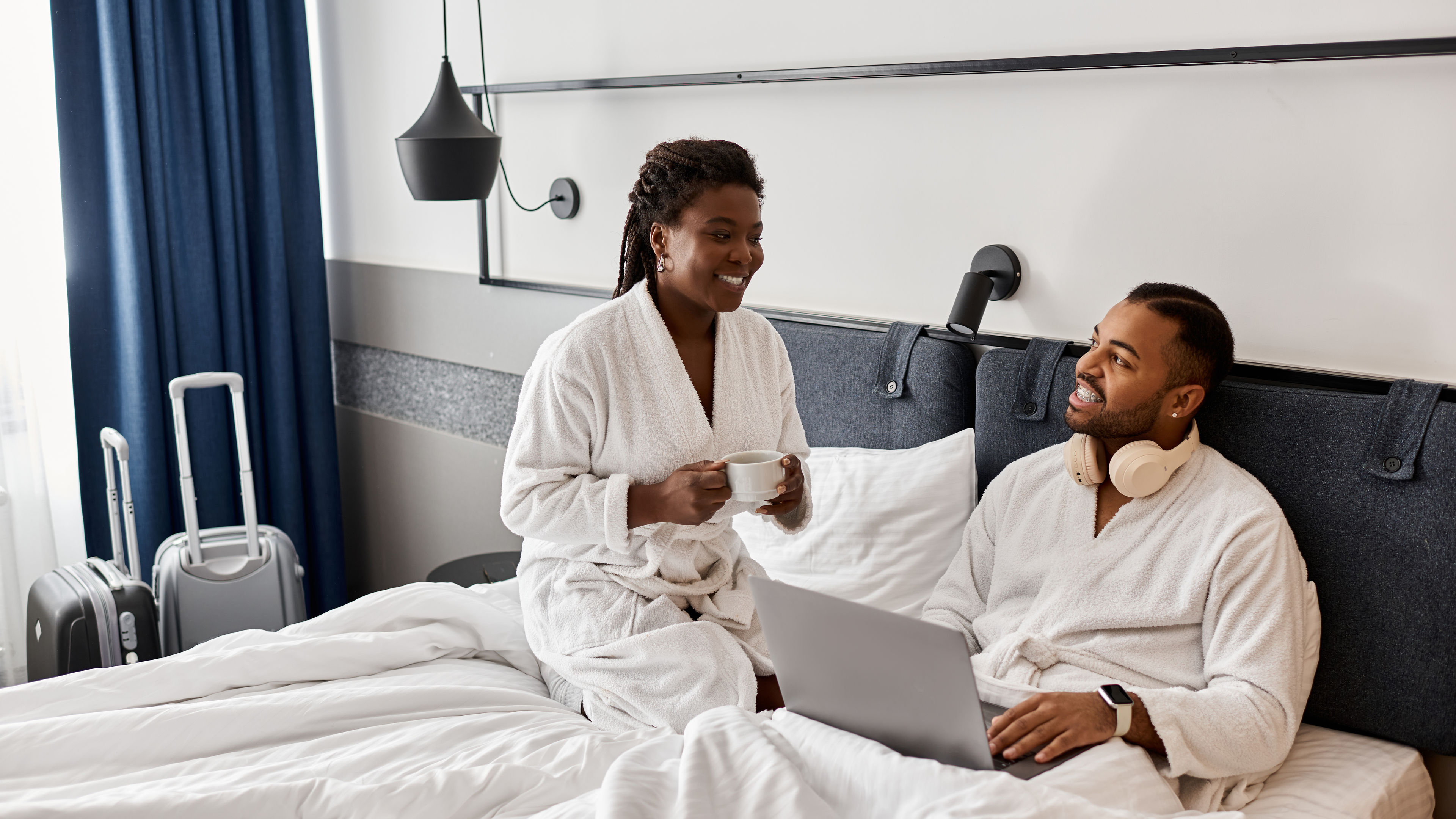 A couple in bathrobes on a bed, one with coffee, the other using a laptop, with suitcases nearby.
