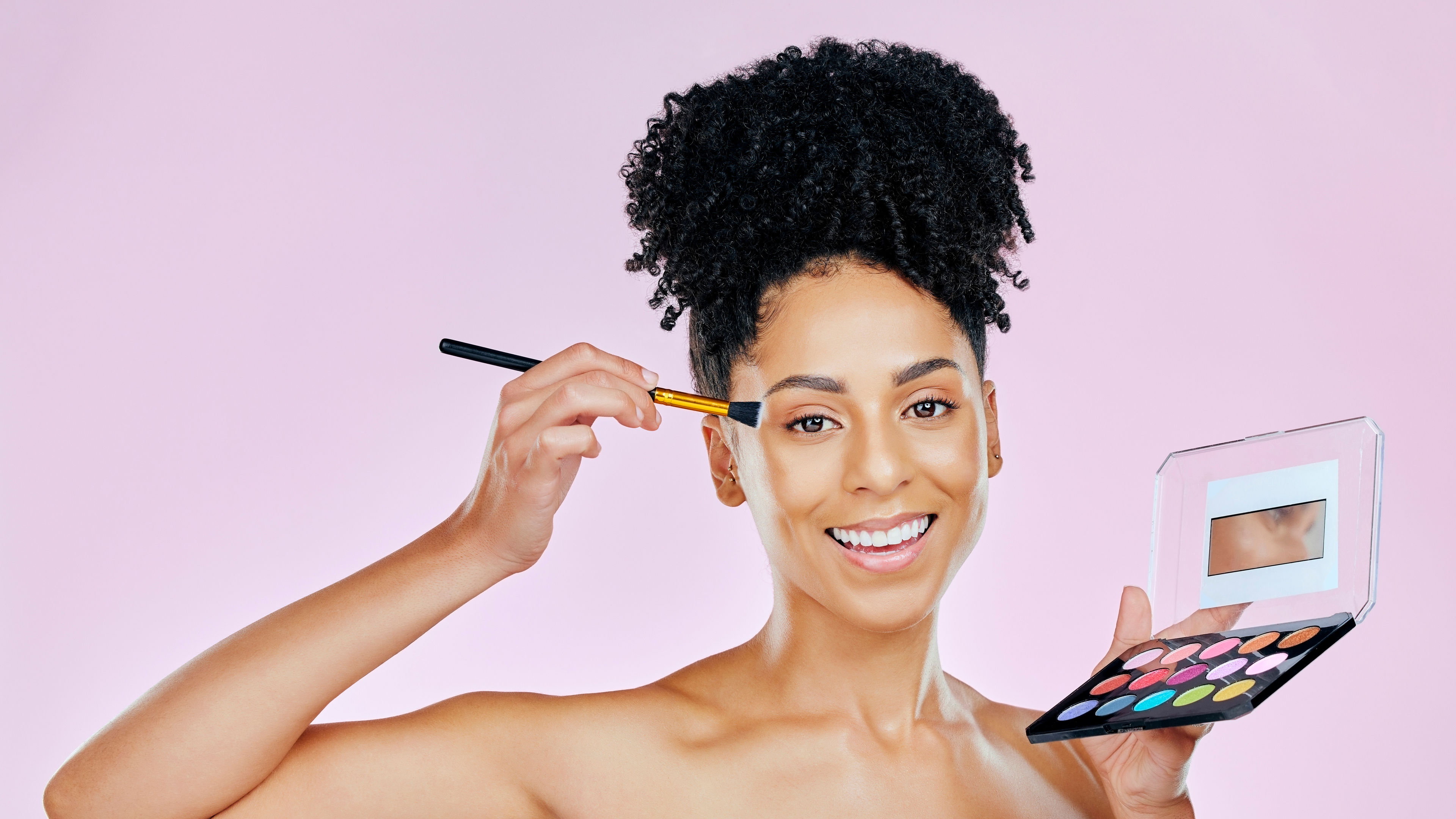 A young woman smiling while applying face powder with a brush, set against a pink background.