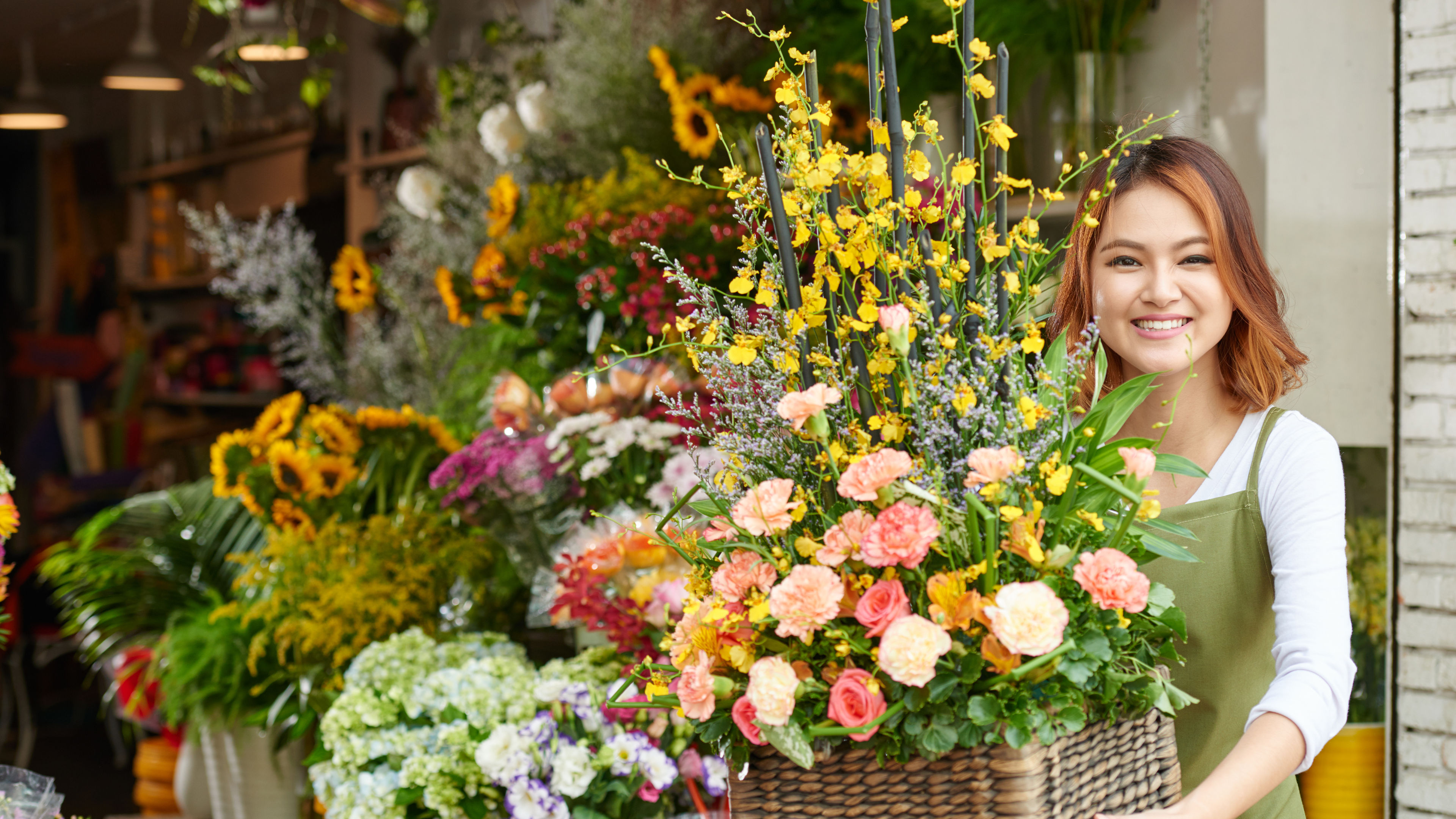 A woman stands in front of a flower shop, holding a basket of flowers.